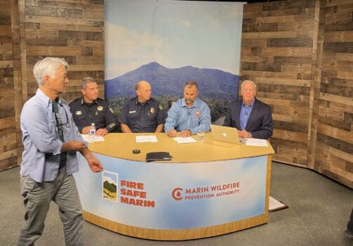 4 men in firefighters dress uniforms sit at a news anchor style desk as a man in casual clothing walks in front of the desk.